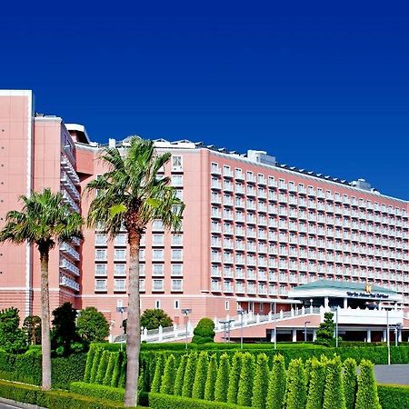 Hotel Grand Nikko Tokyo Bay Maihama Urayasu Esterno foto The image shows a large, multi-story hotel building featuring a light pink facade. The architecture appears modern, with numerous windows arranged in a grid pattern. In the foreground, there are neatly trimmed shrubs and palm trees, adding a tropical