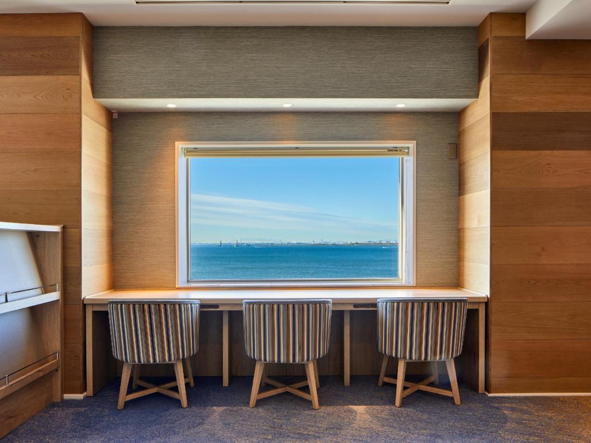 Hotel Grand Nikko Tokyo Bay Maihama Urayasu Esterno foto The photo shows a modern workspace with three striped chairs positioned at a long, white desk. The desk faces a large window with a view of a tranquil body of water and a distant skyline. The walls around the workspace are made of wood paneling, crea