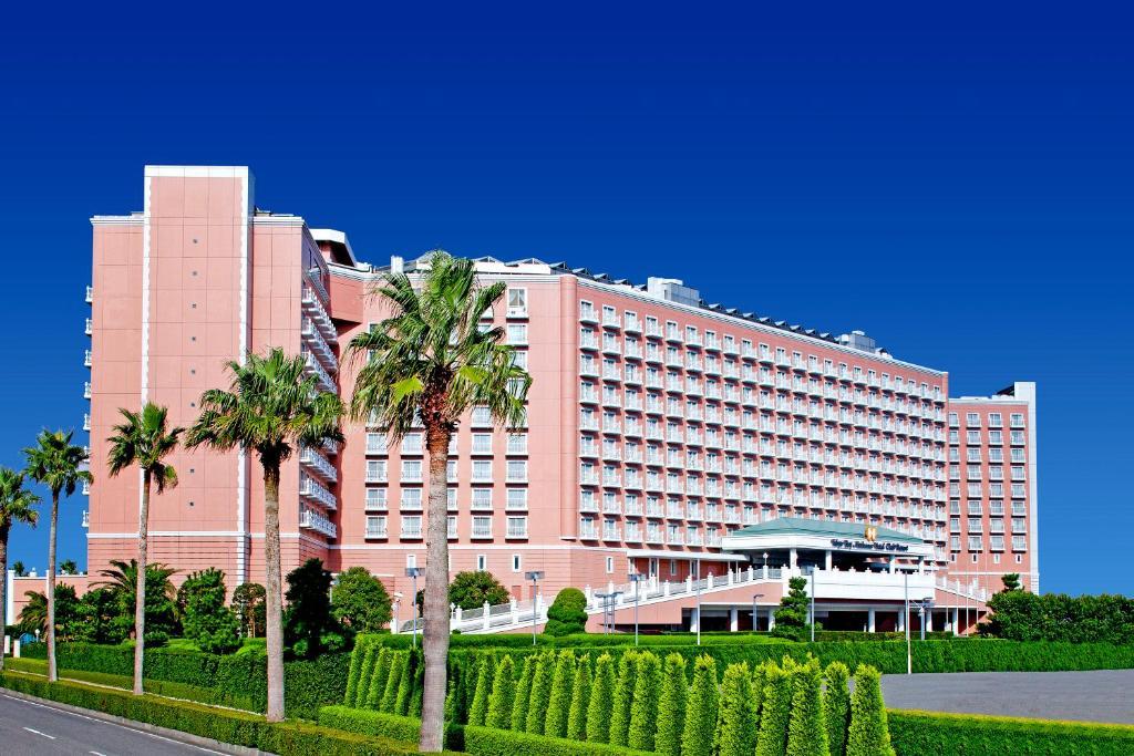 Hotel Grand Nikko Tokyo Bay Maihama Urayasu Esterno foto The image shows a large, multi-story hotel building featuring a light pink facade. The architecture appears modern, with numerous windows arranged in a grid pattern. In the foreground, there are neatly trimmed shrubs and palm trees, adding a tropical